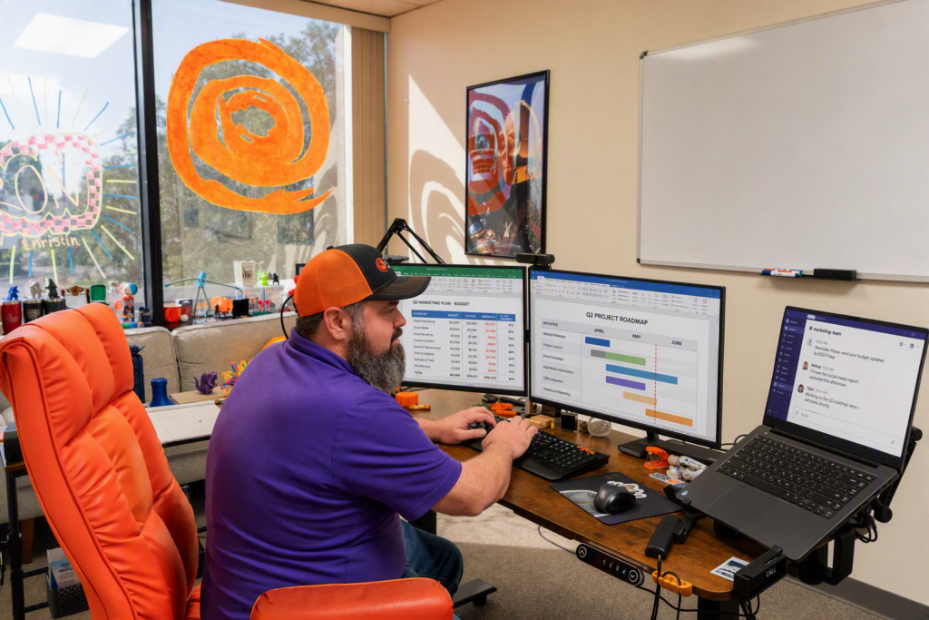 Modern IT professional working at a dual-monitor workstation displaying project management dashboards and business reports in a technology office.